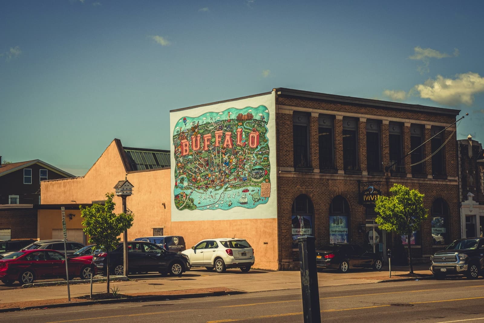 Una calle soleada de Buffalo con un mural colorido del nombre 'BUFFALO' en un edificio de ladrillo.